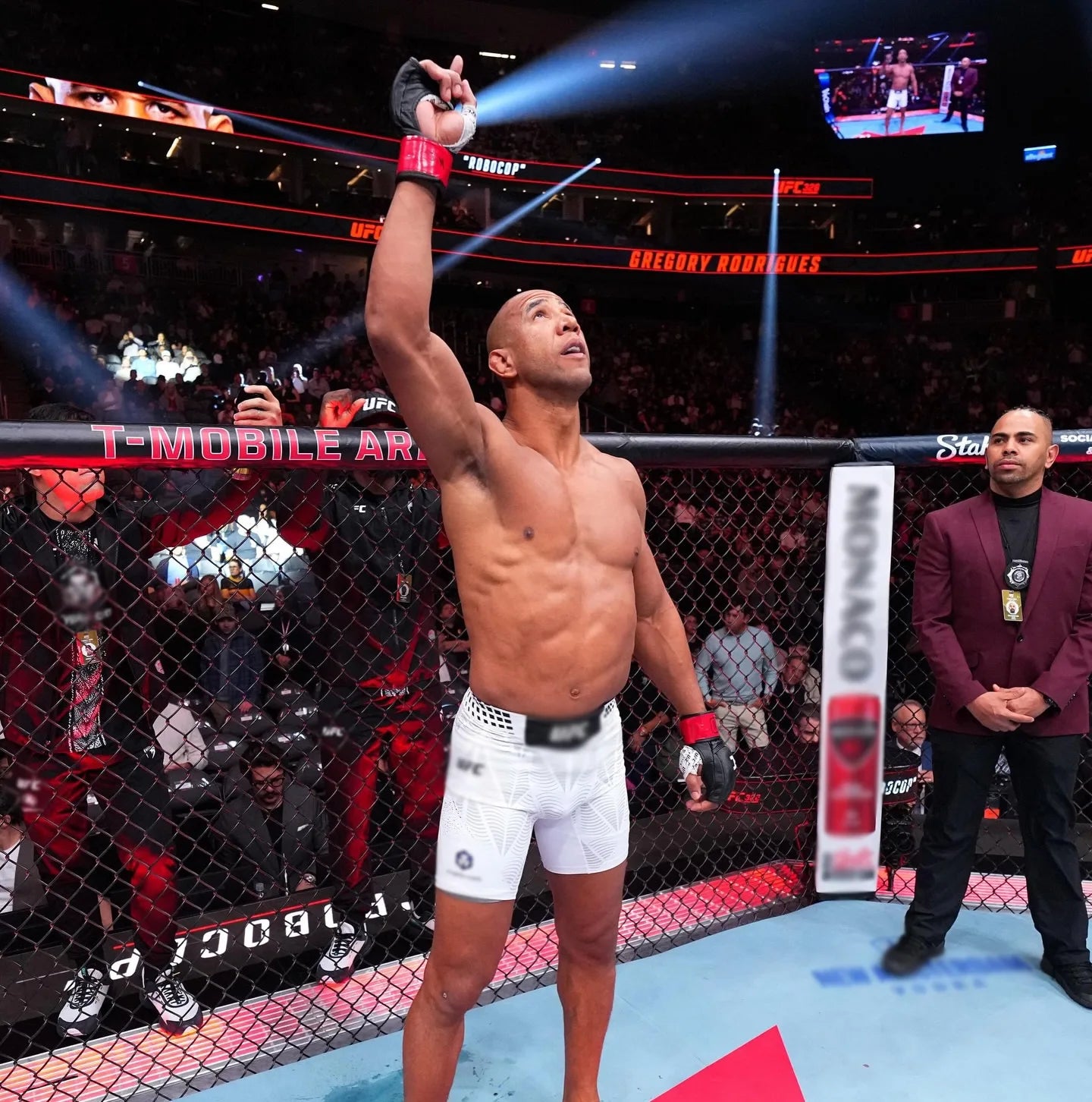 Gregory Rodrigues standing in the octagon with one arm raised toward the lights, the arena crowd behind him
